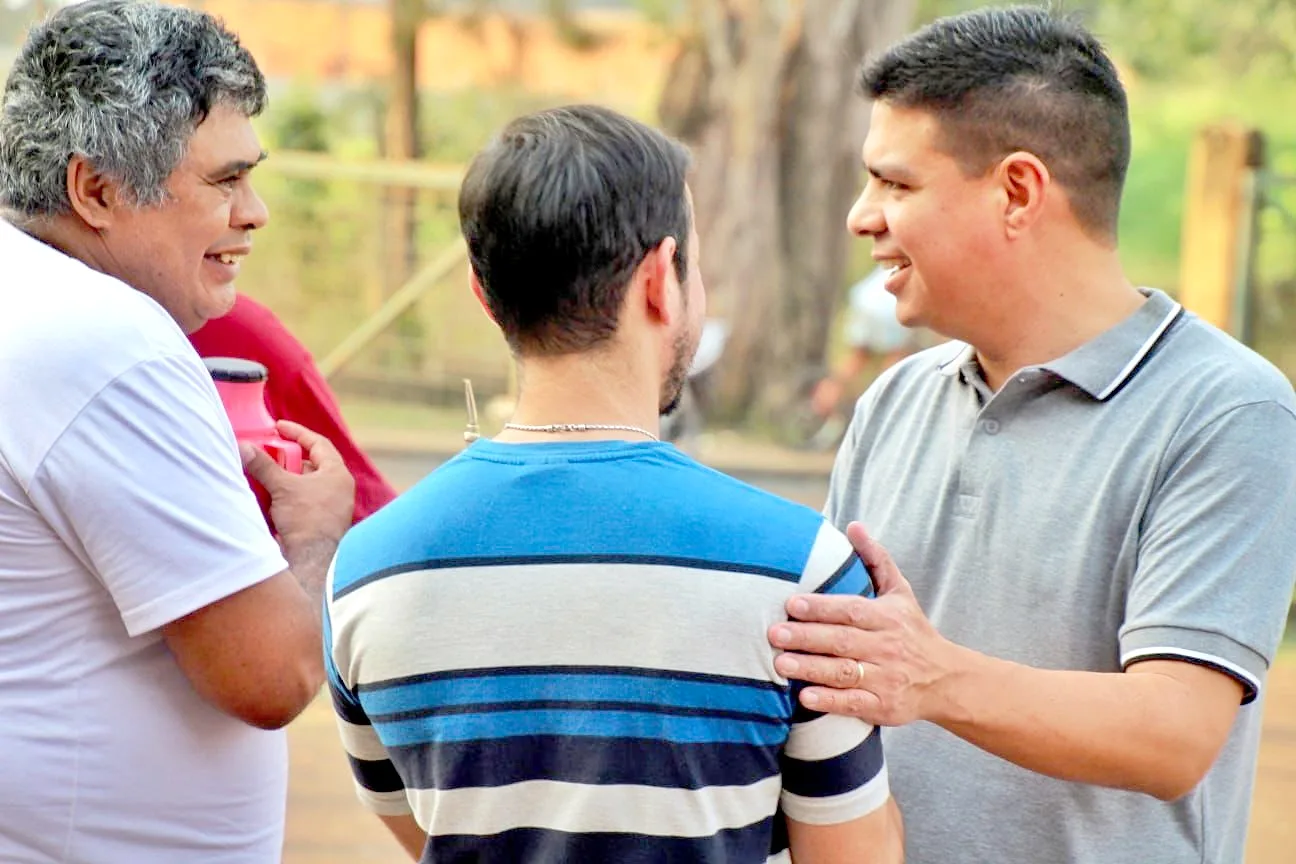 Fernando Meza charla con dos vecinos bajo los árboles, con una mano en el hombro de uno de ellos